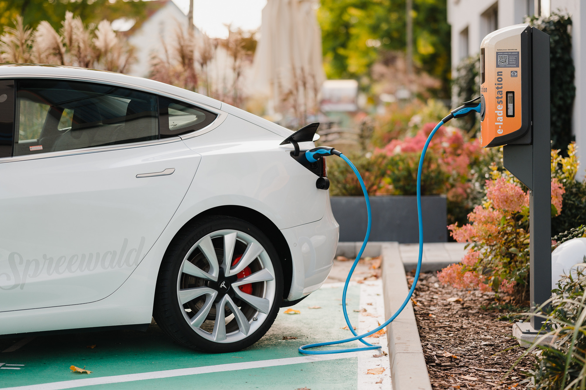 Weißes E-Auto lädt an Ladesäule im Grünen, Kabel am Heck angeschlossen, Station rechts im Bild sichtbar.