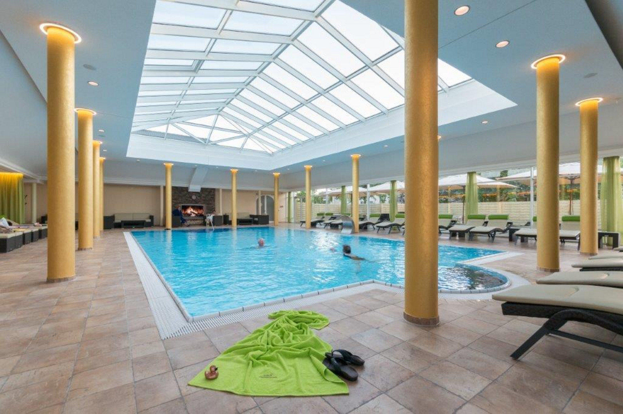 Innenpool mit Glasdach und -wänden, zwei Personen im Wasser, Liegestühle rundherum und grüne Landschaft im Hintergrund.